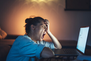 Young woman yawning in front of laptop screen while working from home at night. Deadline and overworking