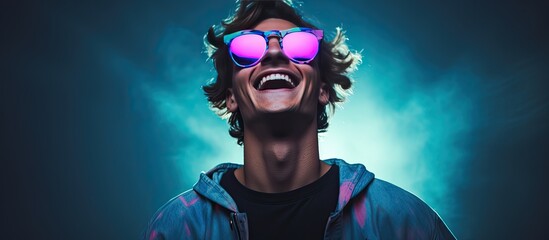 A fashionable teenage boy wearing a black longsleeve and glasses poses with a smile against a blue background illuminated by neon lights representing a hi