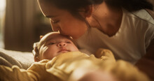 Portrait Of Affectionate Asian Mother Leaning Down To Kiss Her Baby Who Is Resting On A Bed. Woman Expressing Her Motherly Love To Her Infant, Thankful For The Blessing Of Having Beautiful Children.