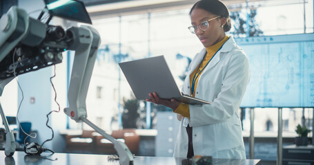 Leinwandbilder - Portrait of Young Black Female Specialist in Lab Coat Using Laptop Computer to Test an AI Robotic Prototype. Professional, Successful Woman Working as an Engineer in High Tech Company Startup