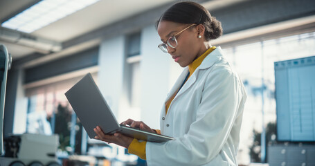 Leinwandbilder - Portrait of Young Black Female Specialist in Lab Coat Using Laptop Computer to Test an AI Robotic Prototype. Professional, Successful Woman Working as an Engineer in Modern High Tech Company Startup