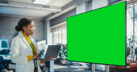 Fototapete - Portrait of Young Black Woman Working in a Lab of Industrial Startup. Female Specialist Using a Laptop Computer and Looking at a Green Mock Up Displayed on Big Digital Screen. Screen Replacement