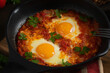 Shakshuka from two eggs in tomato sauce with fresh tomatoes, spices and herbs in a black frying pan. Close-up scrambled eggs