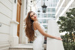 © alones - Glamorous beautiful woman model with brunet hairstyle in fashionable elegant white dress stands and poses near a white vintage building on the street