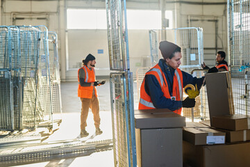 Poster - Young male worker of warehouse sealing one of many cardboard boxes containing metallic spare parts with cellotape against his colleagues