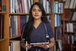 Beautiful Tamil girl with long hair is studying in the library in a medical blue surgical suit with a stethoscope. Doctor, intern, medical student. Back to school