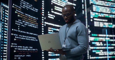 Wall Mural - Portrait of Young Black Man Working on Laptop, Looking at Big Digital Screen Displaying Back-end Code Lines. Professional Programmer Developing a Big Data Interface Software Project Concept.