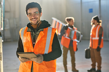 Leinwandbilder - Young successful male technician looking at camera and using tablet while looking through online manual against two colleagues in warehouse