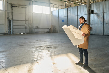 Leinwandbilder - Long shot of young elegant businessman in formalwear holding blueprint with plan of warehouse or some other building where he standing