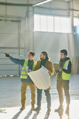 Leinwandbilder - Young confident male engineer pointing at wall of warehouse during discussion of working points and building plan held by contractor