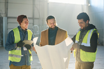 Leinwandbilder - Happy young contractor and two employees in safety jackets looking at sketch or building plan on blueprint while standing in front of camera