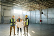 © pressmaster - Three engineers in protective helmets discussing blueprint with building plan while standing in warehouse against sunlight coming through window