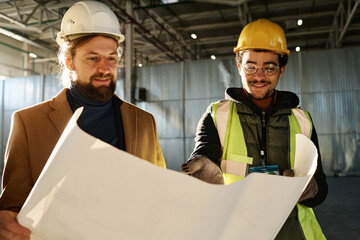 Leinwandbilder - Young smiling engineer explaining details of building plan to contractor or foreman in hardhat while pointing at blueprint at working meeting