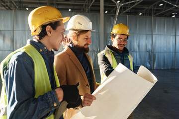Leinwandbilder - Young confident manager in protective helmet and reflective vest explaining sketch on blueprint while standing next to contractor
