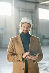 Leinwandbilder - Young successful contractor or foreman in safety helmet and beige coat looking at camera with smile and using tablet against sunlit interior