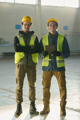 Leinwandbilder - Happy young male colleagues in safety helmets and reflective jackets standing in front of camera in workshop while one of them using tablet