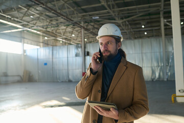 Leinwandbilder - Young confident businessman or contractor in protective hardhat talking on smartphone to his subordinates while standing in warehouse