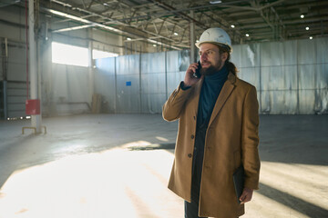 Leinwandbilder - Young successful engineer in casualwear and hardhat talking on mobile phone in workshop while discussing working points with subordinates