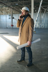 Leinwandbilder - Side view of young bearded engineer in casualwear and protective helmet standing in front of camera in warehouse and speaking on smartphone