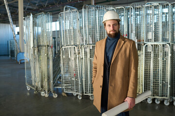 Leinwandbilder - Young modern contractor in hardhat and casualwear holding rolled blueprint in hand and looking at camera while standing in warehouse
