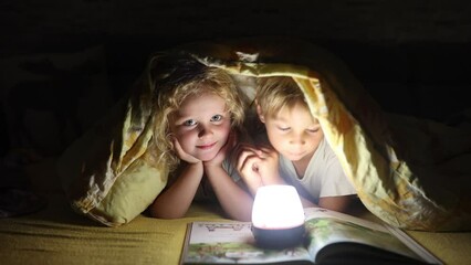 Wall Mural - Two blond cute children, boy and girl, siblings, lying under the cover in bed, reading book together with small light, joy and happiness