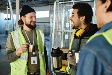 Leinwandbilder - Young cheerful engineer with cup of thermos laughing while talking to colleague with metallic mug with hot tea during break in warehouse