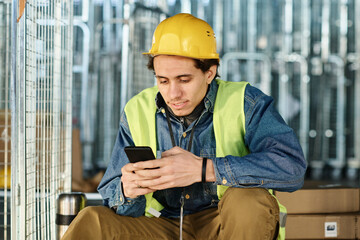 Poster - Young male engineer in workwear and safety helmet texting in smartphone while sitting on large metallic industrial cart with boxes in front of camera