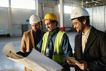 Leinwandbilder - Side view of three young successful architects discussing points of sketch on blueprint held by man in protective helmet and safety jacket