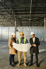 Leinwandbilder - Front view of team of young architects or engineers discussing building plan or sketch on unrolled blueprint held by worker in safety vest