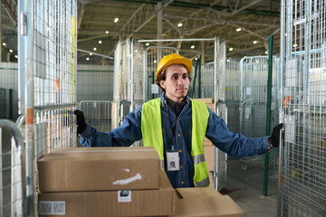 Poster - Young worker of warehouse in safety helmet and reflective vest pushing large metallic cart with stacks of boxes while moving forwards
