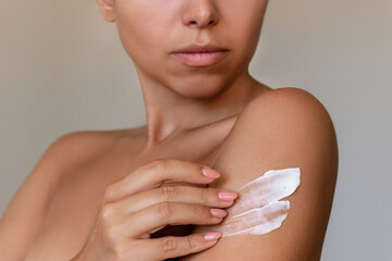 Wall Mural - Cropped shot of young tanned woman moisturizing her shoulder with her hand using a cream isolated on a gray background. Skin care. A white thick smear of cream on the female body. Beauty concept