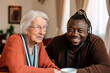 canvas print picture - Junger, afrikanischer Pfleger zuhause bei einer Seniorin 