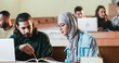 © ihorvsn - Caucasian guy and Arabian girl sitting in auditorium with laptop, talking and working on task at college. Multiethnic male and female at computers discussing studying project at high school.