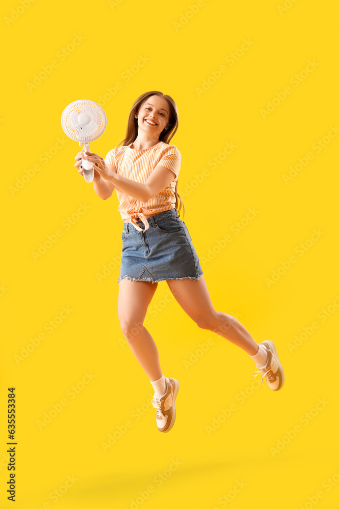 Young woman with electric fan jumping on yellow background