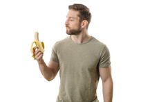 Handsome, Fit Man With A Banana In Hand Eats The Fruit, Chews The Banana, And Looks At The Vegetable While Standing Isolated In A Studio