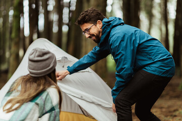 Wall Mural - Cheerful young caucasian couple in jackets in forest enjoy travel vacation, make tent