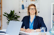 © okrasiuk - Smiling mature businesswoman with laptop at her office workplace, confident middle-aged experienced senior female professional, CEO of company looking at camera. Creative entrepreneur manage business