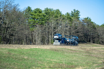 Blue big rig car hauler semi truck transporting vehicles on the modular two level semi trailer driving on the road on the hill with green forest on the background