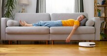Tired Woman Sleeping On Sofa At Home While Robot Vacuum Cleaner Working On Floor