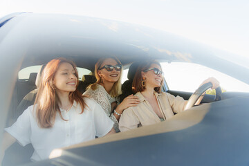 Through car glass front view of young female friends in casual clothes with sunglasses driving in car and looking away while going on summer vacation