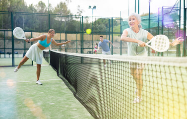 Wall Mural - Senior woman in shorts playing padel tennis on court with young woman. Racket sport training outdoors.