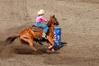 Cowgirl riding in a barrel racing completion at a rodeo. She is wearing red shirt and blue jeans and a white hat. The horse is brown. The barrel is in front of the horse.