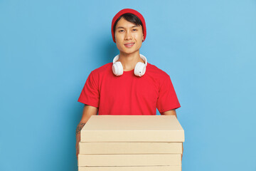 Wall Mural - Smiling delivery guy in red t-shirt with white earphones holds some pizza boxes, standing against blue wall, delisious food concept, copy space