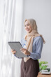 © David - Young business Asian Business muslim woman in purple hijab using tablet and phone, standing near the window in workplace business finance concept.