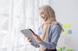 © David - Young business Asian Business muslim woman in purple hijab using tablet and phone, standing near the window in workplace business finance concept.