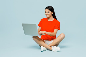 Wall Mural - Full body young IT latin woman she wearing orange red t-shirt casual clothes sitting hold use work on laptop pc computer isolated on plain pastel light blue cyan background studio. Lifestyle concept.