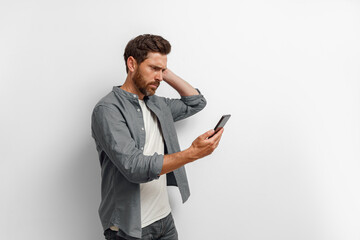 Worried man in casual clothes looking to his mobile phone while standing on white background