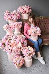 Wall Mural - a happy young girl holds a box of flowers in her hands and inhales the fragrance of flowers. A beautiful woman is sitting on a sofa surrounded by flowers. A box with peonies and bush roses in a peach