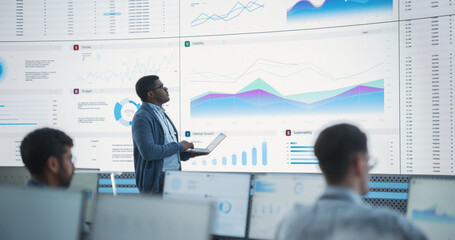 Fototapete - Male Financial Data Analyst Using Laptop And Studying Information And Graphs On Big Digital Screen In Monitoring Office. Black Man Working For Big Consulting Firm, Diverse Employees Behind Computers.