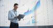 © Gorodenkoff - In The Office: Male Monitoring Specialist Holding Laptop Computer And Standing Next To Big Digital Screen With Graphs And Charts. Caucasian Man Analyzing Data, Performance Records, And Filing Report.